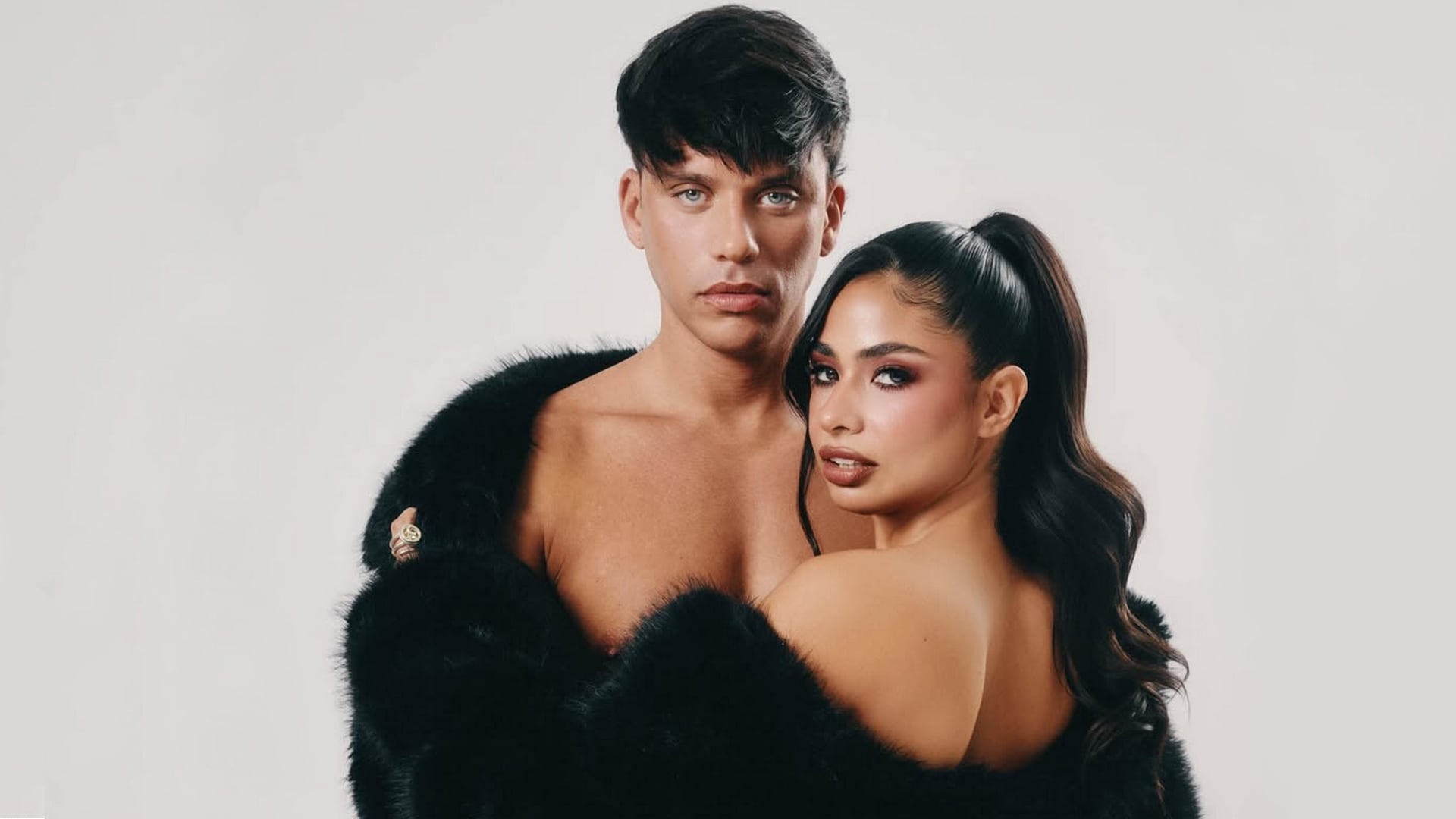 En una sorprendente sesión, Daniela Celis y Nick Sicaro posan sin camisa, cubiertos solo por un elegante abrigo de piel negro sobre un fondo blanco, irradiando confianza y estilo.