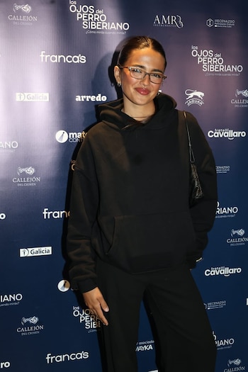 Maia Reficco sonríe con anteojos y sudadera negra con capucha frente a un fondo publicitario con logotipos