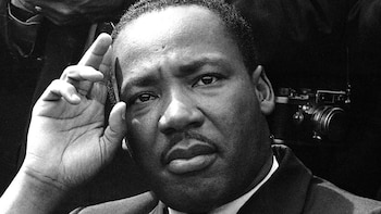 Retrato en blanco y negro de Martin Luther King Jr. con una expresión seria, mirando al frente. Su mano izquierda está levantada, con los dedos tocando su sien derecha