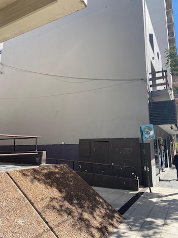 Fachada de edificio gris con rampa de hormigón texturizado, pasamanos metálicos, cartel de accesibilidad y acera con sombras de árboles en un día soleado