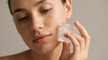 Primer plano del rostro de una persona con pecas, que sostiene un cubo de hielo grande y transparente contra su mejilla derecha. Una gota de agua se desprende.