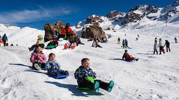 Vista frontal de niños riendo mientras se deslizan en trineos sobre una pendiente nevada. Detrás, más personas caminan y montañas cubiertas de nieve.