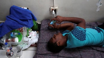 Primer plano de un joven recostado en el suelo, usando un smartphone que está conectado a un enchufe de pared. Al lado hay botellas de plástico y una manta azul