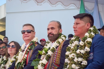 Tres hombres adultos lucen guirnaldas florales blancas y verdes; uno con barba, otro con gafas de sol, y el tercero con traje azul, mirando a la derecha