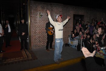 Un hombre con sudadera blanca, gorra y brazos alzados canta ante varias personas que sostienen teléfonos móviles y una bandera argentina