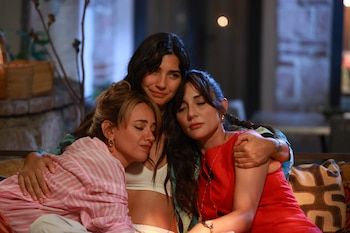 Tres mujeres se abrazan en un sofá. La mujer del centro, con cabello oscuro, sonríe. La mujer de la izquierda, rubia, y la de la derecha, morena, tienen los ojos cerrados