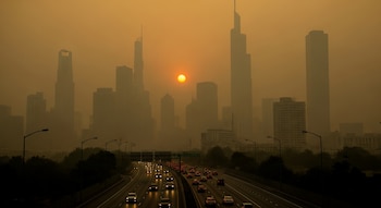 Horizonte urbano con rascacielos sumergidos en neblina naranja. El sol bajo brilla en el centro. Una autopista con tráfico ligero cruza la parte inferior.