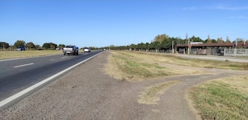 Carretera de asfalto con línea blanca y una vía de tierra paralela a la derecha. Varios autos circulan. Hay árboles a los lados y un cielo azul despejado