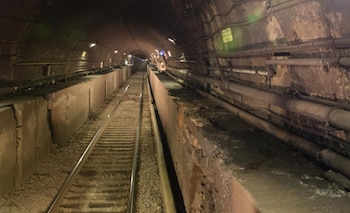 Interior de un túnel ferroviario subterráneo con una vía de tren, paredes de hormigón, tuberías laterales y focos en el techo