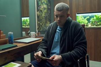 Un hombre de pelo gris vestido con una bata gris y camiseta azul mira unos papeles en un escritorio de madera; se ven un portátil y dos acuarios