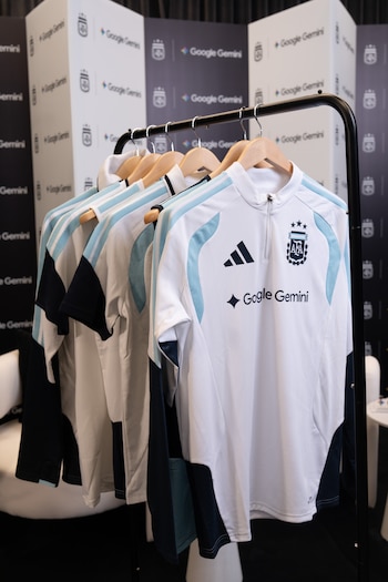 Camisetas de entrenamiento de la Selección Argentina, blancas, celestes y azul oscuro, colgadas en un perchero, con logos de Adidas, AFA y Google Gemini. El fondo tiene patrones del logo Google Gemini
