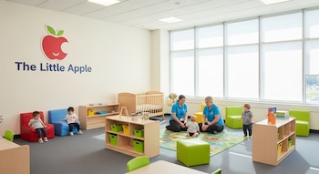 Vista amplia de un aula de guardería luminosa con niños jugando, dos adultos, mobiliario colorido, cunas, y el logo 'The Little Apple' en la pared.