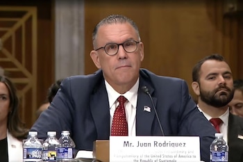Hombre con traje, corbata roja, gafas, micrófono y pin bandera de Estados Unidos. Cartel con su nombre. Botellas de agua. Otras personas en segundo plano