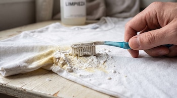 Mano aplica pasta blanca con un cepillo sobre la mancha de una camiseta blanca colocada en una mesa.