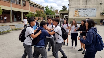 Un grupo de alumnos con uniforme escolar forcejea en un patio pavimentado frente a un cartel del Colegio Secundario San José.