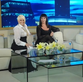 Tres mujeres, Carmen Barbieri, Moria Casán y Susana Giménez, sentadas en un sofá blanco frente a una mesa de vidrio con flores y copas