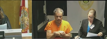 Un hombre con uniforme naranja y esposas permanece sentado al lado de una mujer con documentos y la bandera de Florida