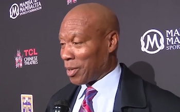 Byron Scott habla ante un micrófono con traje oscuro y corbata frente a un fondo con logotipos