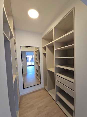 Vestidor vacío con estanterías y cajones de madera clara y suelo de parqué, con un espejo de cuerpo entero que refleja una habitación luminosa