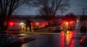 Noche en pueblo de EE. UU. Calle mojada iluminada por luces rojas y azules de equipos Hazmat, bomberos y policía rodeando una casa acordonada.