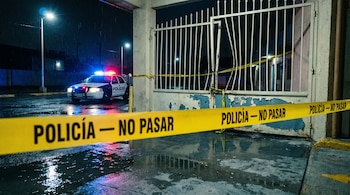 Cinta amarilla de 'Policía — No Pasar' en primer plano frente a un portón blanco de motel dañado bajo la lluvia nocturna. Una patrulla ilumina el fondo con luces.