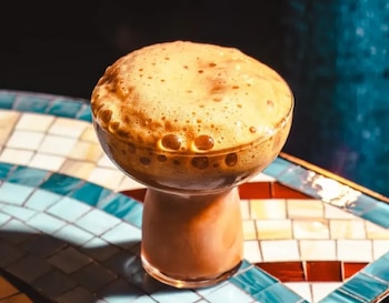 Primer plano de un cóctel oscuro con mucha espuma de color canela en un vaso de cristal con pie corto, sobre una mesa de mosaico con azulejos azules, blancos y rojos