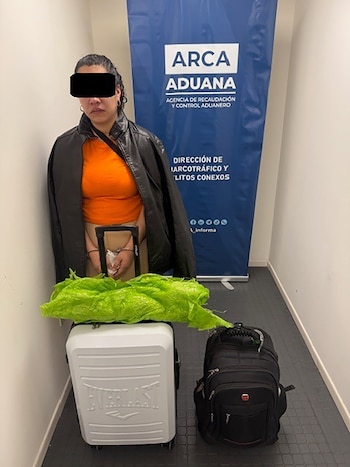 Una mujer, con rostro censurado y manos esposadas, frente a un cartel azul de Aduana, con una maleta blanca, una mochila negra y un bulto verde