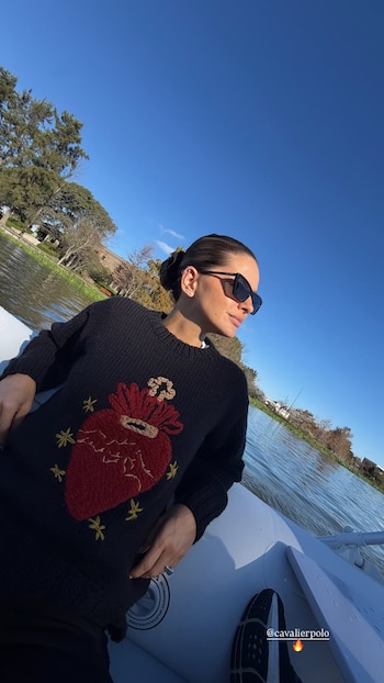 Mujer con suéter oscuro bordado, lentes de sol y cabello recogido sentada en un bote blanco sobre el agua con cielo azul y árboles de fondo