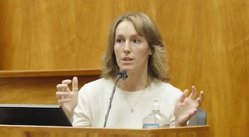Una mujer de cabello castaño claro y blusa clara habla ante un micrófono, gesticulando con ambas manos en un entorno con paneles de madera clara