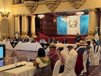 Salón ornamentado con columnas y candelabros. Personas en mesas y audiencia asisten a una reunión, con banderas de Guatemala al fondo