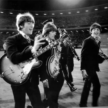 Fotografía en blanco y negro de John Lennon y Paul McCartney de The Beatles, caminando en un estadio con sus guitarras y trajes