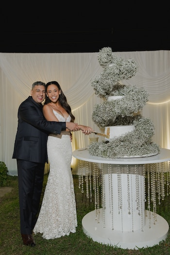Una mujer vestida de novia y un hombre en traje oscuro cortan un pastel de bodas decorado con flores sobre una mesa redonda
