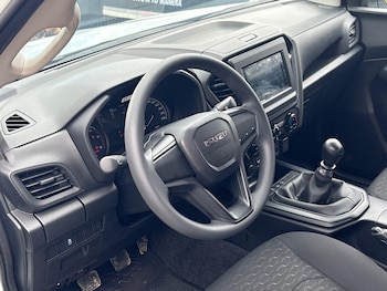 Interior de un vehículo con volante negro Isuzu, tablero, pantalla de infoentretenimiento, palanca de cambios manual y pedales del conductor
