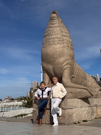 Una pareja posa frente a una gran escultura de piedra de un lobo marino con la boca abierta. Al fondo se ven edificios, un puerto y un cielo azul claro