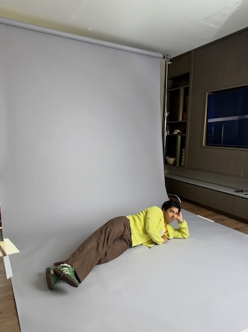 Joe Jonas, de cabello oscuro y rizado, yace de lado en un fondo gris, vestido con un suéter amarillo y pantalones marrones