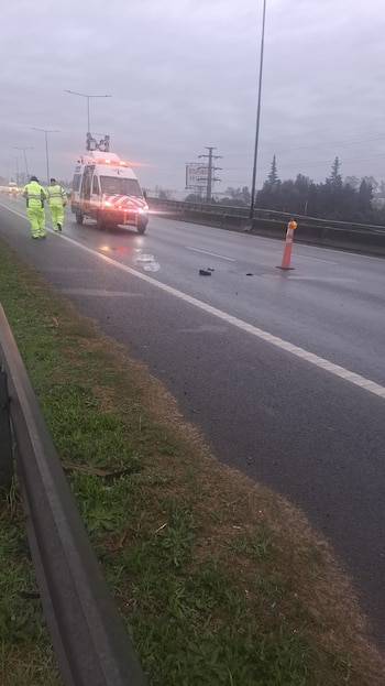 Carretera mojada, vehículo de servicio con luces intermitentes, tres personas con chalecos reflectantes, cono de tráfico, escombros en el asfalto, valla metálica