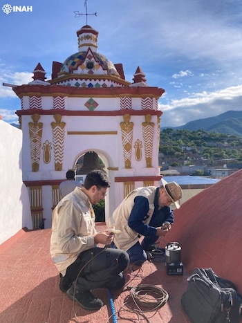 Dos hombres en cuclillas en una azotea roja. Manipulan cables y un dispositivo junto a un campanario blanco con decoración. Montañas verdes al fondo