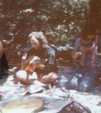 Imagen descolorida de Indio Solari sentado con las piernas cruzadas en el suelo, tocando una guitarra acústica, con árboles de fondo y otra persona