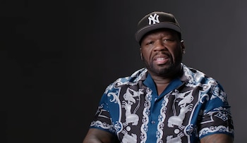 50 Cent con gorra de béisbol negra con el logo de los Yankees, camisa estampada azul y blanca, y pendientes, posa de frente sobre un fondo gris oscuro