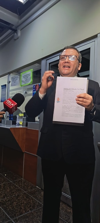 Un hombre con traje y gafas sostiene un documento frente a una ventanilla de la Corte de Constitucionalidad y habla ante un micrófono de prensa