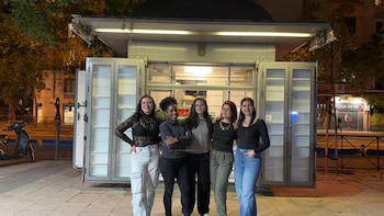 Cinco mujeres sonrientes posan de noche frente a un moderno kiosco con paneles de vidrio iluminados. Al fondo se ven árboles y una calle urbana