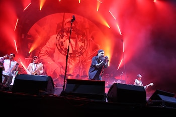 Una banda de rock actúa en un escenario oscuro bañado en luces rojas. Se ve al vocalista, trombonista, saxofonista y guitarrista tocando
