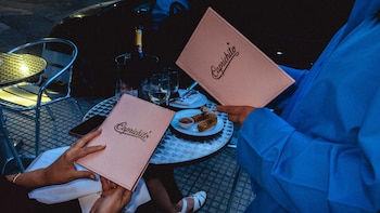 Primer plano de dos personas sosteniendo menús rosados de 'Caprichito' en una mesa exterior con un plato de frituras, copas de vino y champán