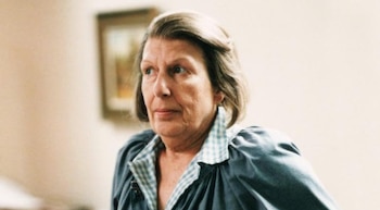 Retrato de una mujer mayor con cabello corto y gris, con una blusa azul sobre una camisa a cuadros azul y blanco, mirando hacia la izquierda en un fondo claro