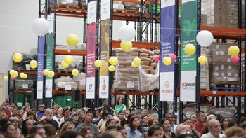 Vista de una multitud sentada durante un evento en un almacén, con estanterías altas llenas de cajas y sacos, y linternas decorativas colgando