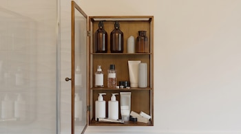 Gabinete de baño de madera con puerta de cristal abierta, revelando estantes con botellas de bomba, tubos y recipientes en tonos neutros. Pared clara de fondo.