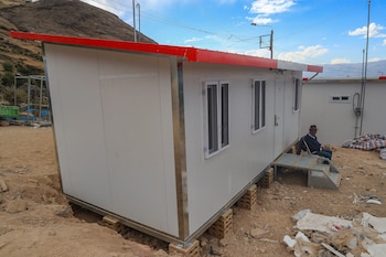 Un módulo de vivienda prefabricado de color blanco y borde rojo instalado sobre tierra, con un hombre sentado a un lado