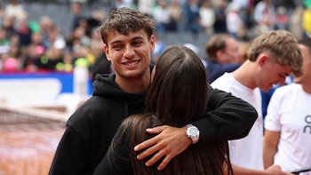 Primer plano de Flavio Cobolli sonriendo y abrazando a una mujer de espaldas, con cabello oscuro. Él lleva una sudadera negra y reloj de pulsera