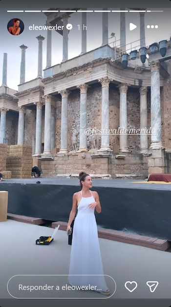 Una mujer con vestido blanco está en un escenario con columnas de un teatro romano al fondo. Hay fardos de heno y una caja de herramientas en el suelo