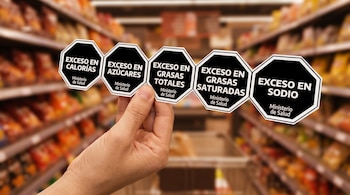 Una mano sostiene cinco sellos octagonales negros con bordes blancos y texto blanco. Se lee "Exceso en Calorías", "Exceso en Azúcares", "Exceso en Grasas Totales", "Exceso en Grasas Saturadas", "Exceso en Sodio", y "Ministerio de Salud". El fondo muestra estantes de supermercado.